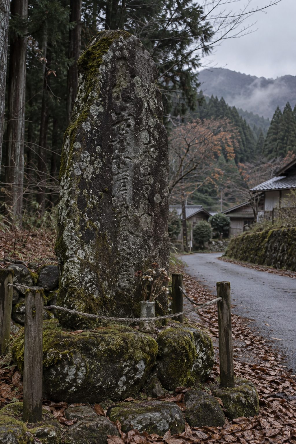 旧街道沿いに立つ、苔むした縦長の石の慰霊碑。周りに縄が張られている。